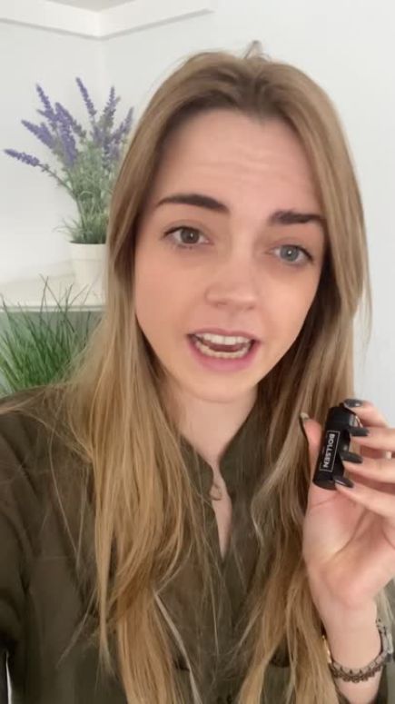 A woman with long blonde hair holds a black nail polish bottle, surrounded by greenery.