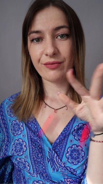 Woman in a blue patterned top, gesturing with pink objects against a grey background.