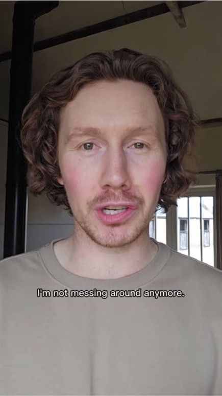 A man with curly hair wearing a beige shirt, speaking in a bright room.