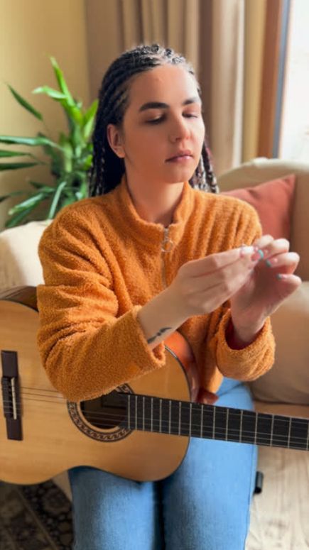 Woman in an orange sweater holds a small object, sitting beside a guitar.