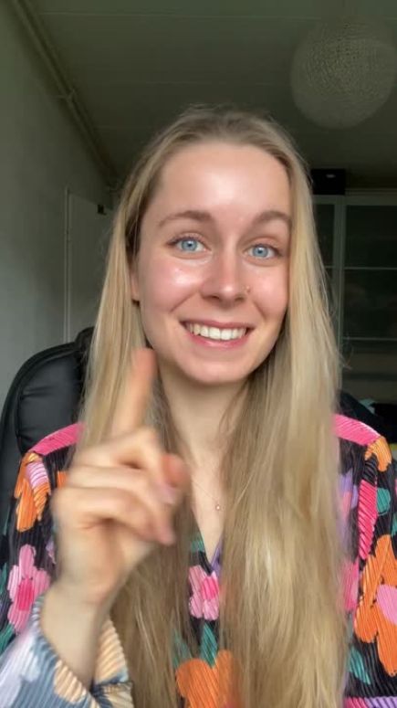 Smiling woman with long blonde hair, wearing a colourful floral blouse, gesturing with her finger.