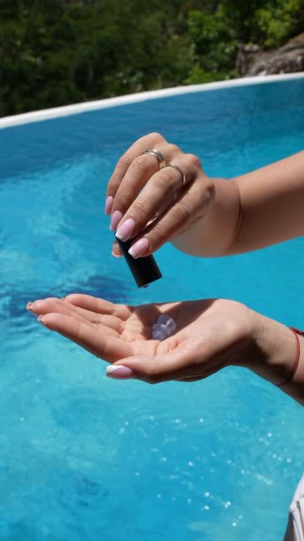 A hand with pink nails holds a black bottle over a palm, near a blue pool.