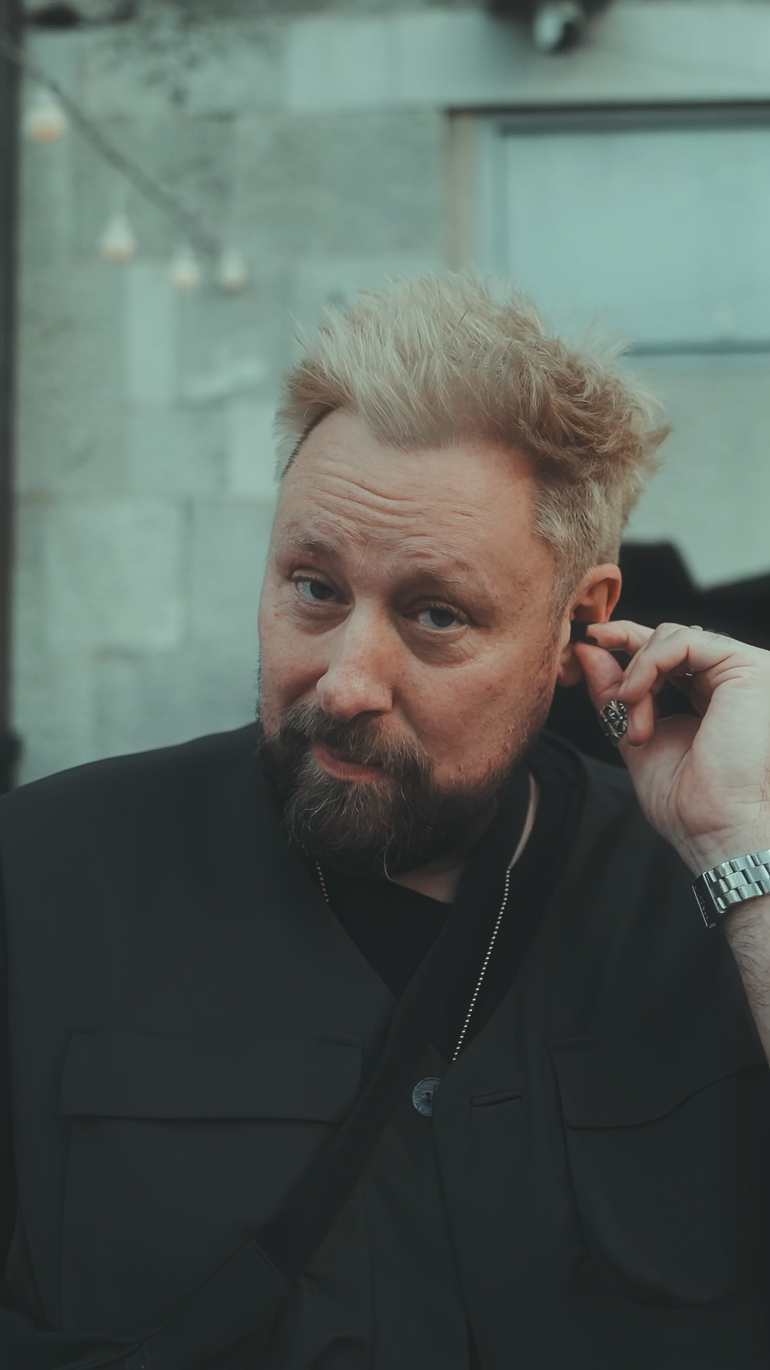 Man with blonde hair and beard, wearing a black jacket, touching his ear.