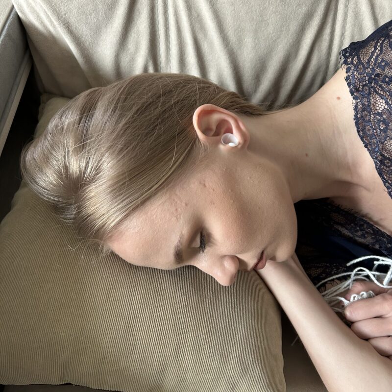 Sleeping woman with blonde hair resting on a beige cushion, wearing white earbuds.