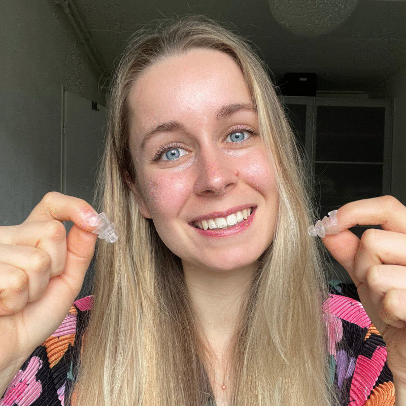 Smiling woman with long blonde hair holding clear earplugs, wearing a colourful patterned top.