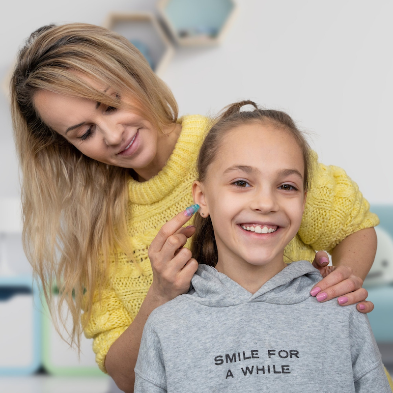 A woman in a yellow sweater fits BOLLSEN Earplugs Hearing Protection Kidz+ on a smiling girl.
